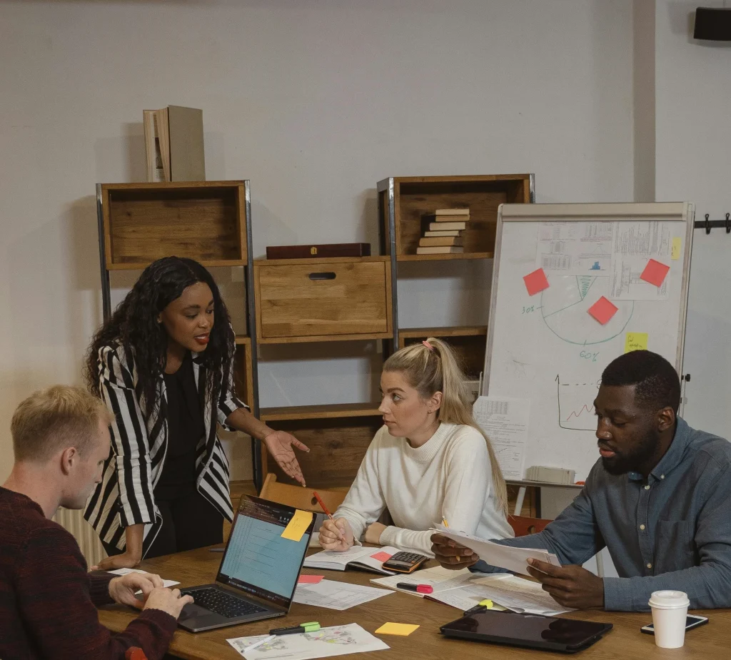 A meeting around a table with four people reviewing documents and a website design. A laptop, a whiteboard, and other planning materials are visible.